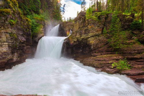A serene waterfall cascades over rocky cliffs, surrounded by lush greenery and trees under a bright blue sky.