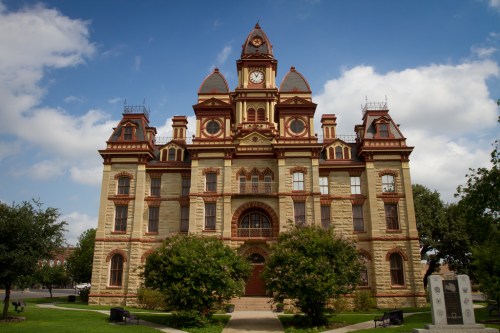 Historic building with a clock tower, ornate architecture, and lush greenery in front under a blue sky.