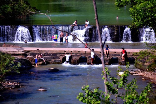 People enjoying a sunny day by a waterfall, playing in the water and relaxing along the riverbank.