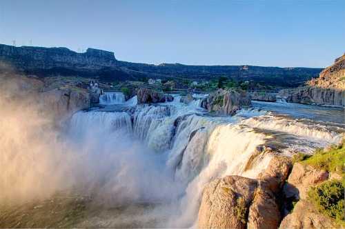 A stunning waterfall cascades over rocky cliffs, surrounded by lush greenery and a clear blue sky.