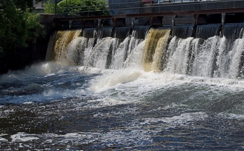 A waterfall cascades over a dam, creating white foam in the river below, surrounded by greenery and a clear blue sky.