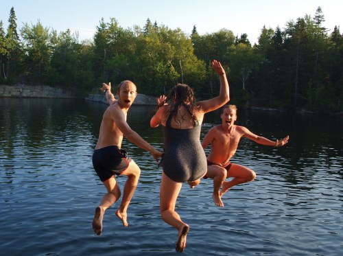 Three people joyfully jumping into a lake on a sunny day, surrounded by trees.