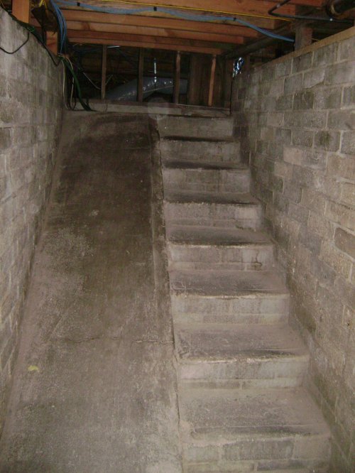 A dimly lit basement with a concrete ramp on the left and a set of stairs on the right, both leading upward.