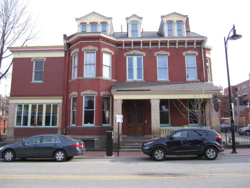 A large red brick building with multiple windows and a decorative roof, flanked by parked cars on a street.