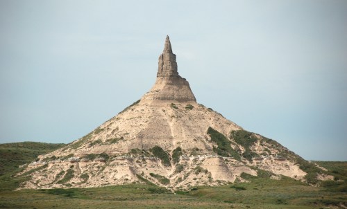 A tall, pointed rock formation rises from a grassy landscape under a clear sky.