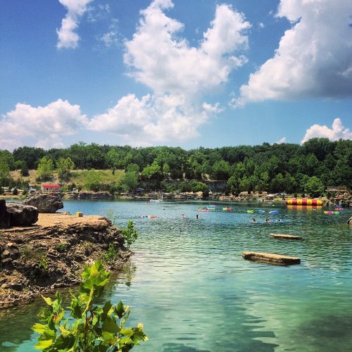 A scenic lake surrounded by greenery, with people swimming and colorful inflatables in the water under a blue sky.