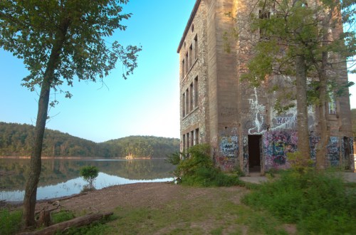 Abandoned building by a calm lake, surrounded by trees and hills, with colorful graffiti on its walls.