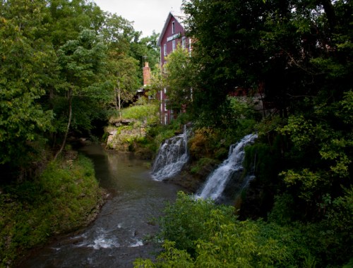 A serene scene featuring a flowing creek with small waterfalls, surrounded by lush greenery and a rustic building in the background.