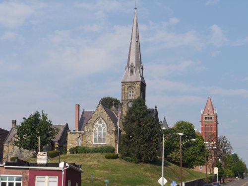 A historic church with a tall steeple sits atop a grassy hill, surrounded by trees and buildings under a clear sky.