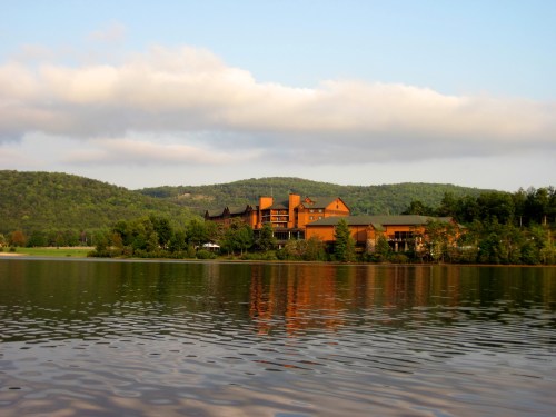 A serene lakeside view of a lodge surrounded by green hills under a partly cloudy sky. Reflections shimmer on the water.