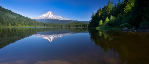 A serene lake reflects a snow-capped mountain surrounded by lush green forests under a clear blue sky.
