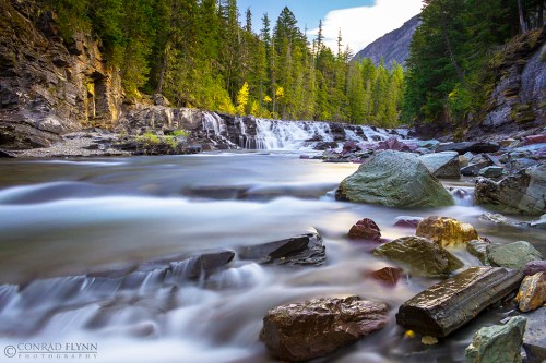 A serene river flows over rocks, surrounded by lush green trees and mountains under a clear blue sky.