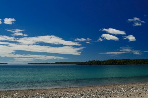 A serene beach scene with clear blue water, pebbly shore, and a backdrop of green trees under a bright sky.