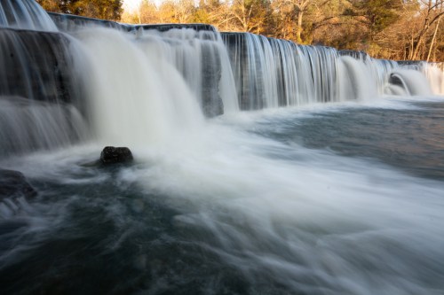A serene waterfall cascades over rocks into a calm pool, surrounded by trees in a tranquil natural setting.