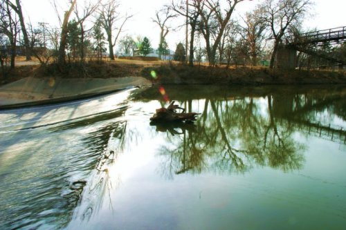 A serene river scene with trees reflecting in the water and a bridge in the background, illuminated by soft sunlight.