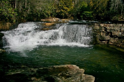 A serene waterfall cascades over rocky ledges into a clear pool, surrounded by lush greenery and trees.