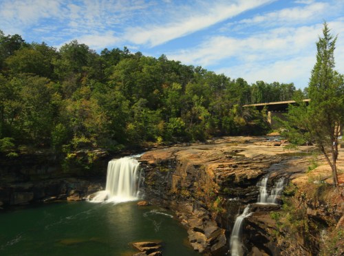 A serene waterfall cascades into a green pool, surrounded by rocky cliffs and lush trees under a blue sky.