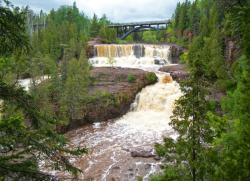 A scenic view of a waterfall surrounded by lush greenery and a bridge overhead, with rushing brown water below.
