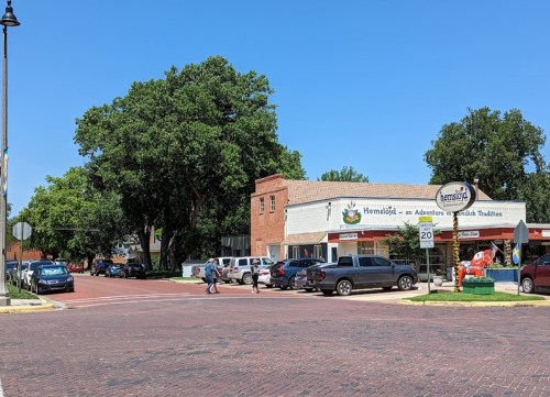 A busy street corner with shops, parked cars, and pedestrians crossing on a sunny day. Green trees in the background.