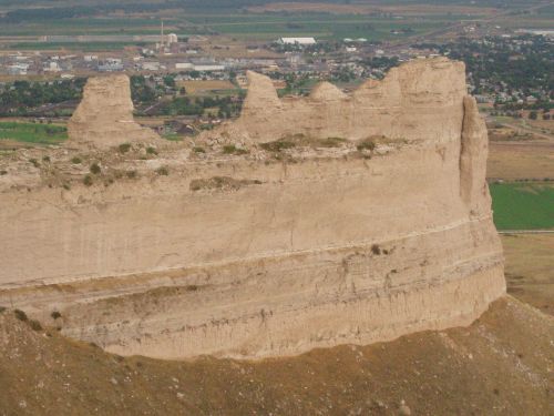 A rugged cliff formation overlooking a valley with fields and a small town in the distance.
