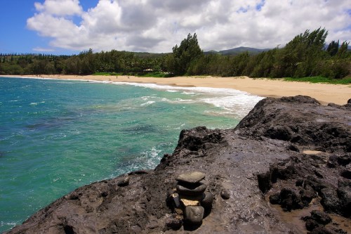 A rocky shoreline with stacked stones, turquoise water, and a sandy beach under a partly cloudy sky.