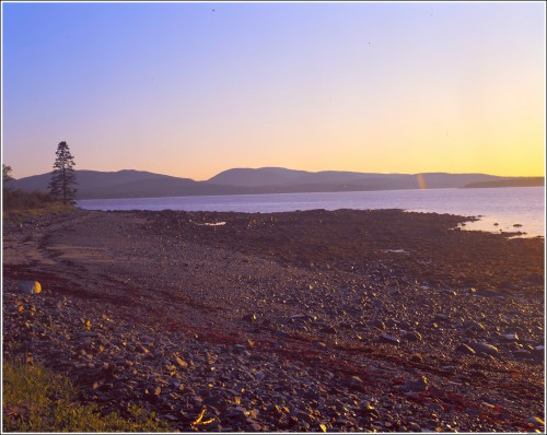 A serene beach at sunset, featuring rocky shorelines and distant hills under a colorful sky.