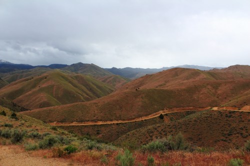 Rolling hills with green and brown terrain under a cloudy sky, featuring a winding dirt road through the landscape.