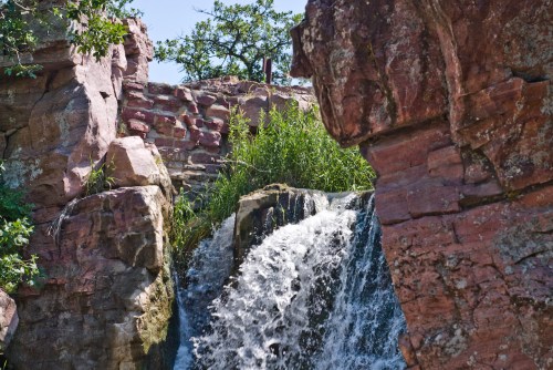A waterfall cascades over rocky cliffs, surrounded by lush greenery and a clear blue sky.