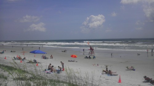 A sunny beach scene with people relaxing, colorful umbrellas, and a lifeguard tower near the ocean waves.
