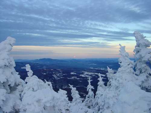 A snowy landscape with frosted trees, overlooking a valley at sunset under a cloudy sky.