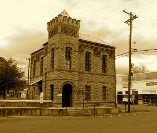 Historic brick building with a turret, set in a small town, surrounded by power lines and shops. Sepia-toned image.
