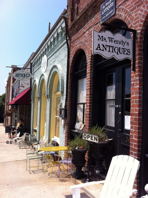 Quaint street view of antique shops with colorful facades and outdoor seating, featuring "Ms. Wendy's Antiques" sign.