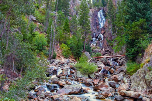 A serene waterfall cascades down rocky terrain, surrounded by lush green trees and a flowing stream below.