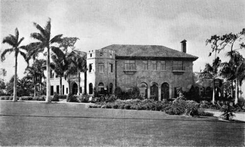 Historic two-story house with arched windows, a tile roof, and palm trees in the foreground.