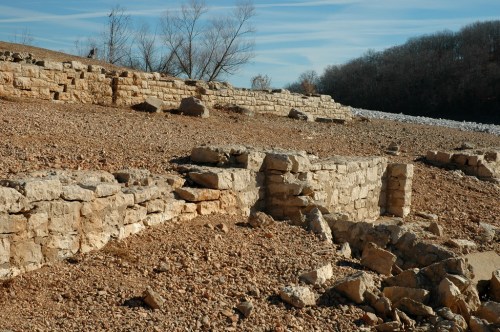 Ruins of stone walls and foundations on a dry, rocky landscape with sparse trees in the background.