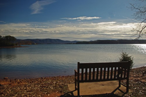 A wooden bench by a tranquil lake, with mountains and a cloudy sky in the background.
