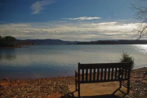 A wooden bench by a tranquil lake, with mountains and a cloudy sky in the background.