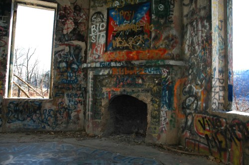 Graffiti-covered walls surround a dilapidated fireplace in an abandoned building with windows revealing a wooded view.