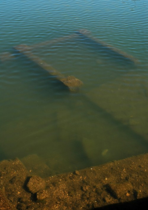 Submerged wooden structure partially visible in clear water, with rocks and sediment on the bottom.