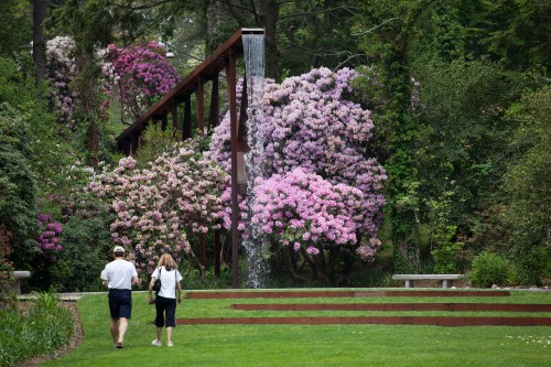 Two people walk through a lush garden with vibrant pink flowers and a waterfall in the background.