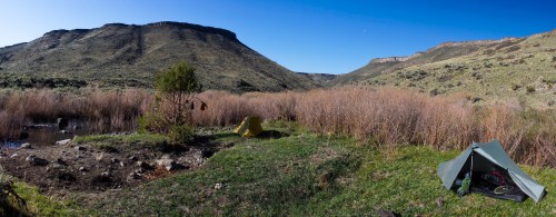 A scenic landscape featuring a tent by a pond, surrounded by tall grass and mountains under a clear blue sky.