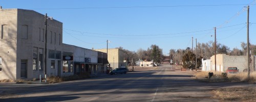 A quiet, empty street in a small town with storefronts and sparse vegetation under a clear blue sky.