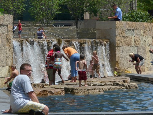 Children play in a water feature with a waterfall, while adults supervise and relax nearby on a sunny day.