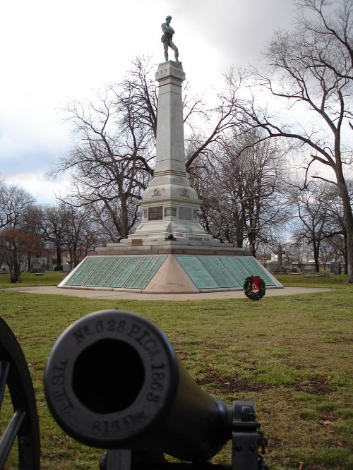A war monument with a statue atop a tall pedestal, surrounded by trees and a cannon in the foreground.