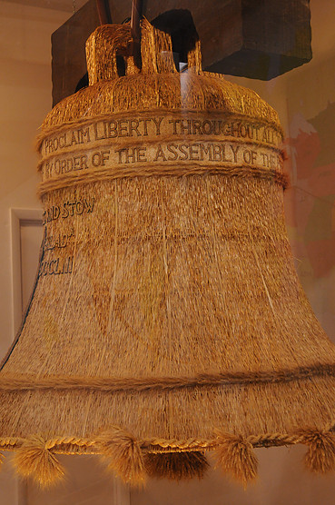 A large, ornate bell with inscriptions, displayed in a glass case, featuring a straw-like texture.