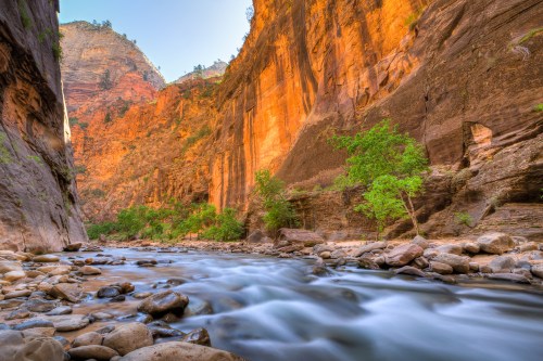 A serene river flows through a narrow canyon, surrounded by towering orange cliffs and lush greenery.