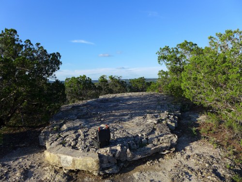 A flat stone platform surrounded by greenery under a clear blue sky, with distant hills visible in the background.