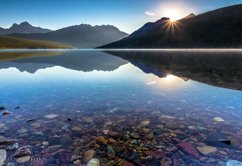A serene lake at sunrise, reflecting mountains and colorful stones beneath the clear water.