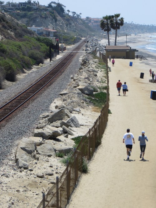 A sandy path along the coast with people walking, train tracks nearby, and palm trees in the background.