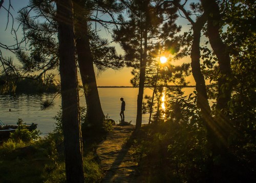 A silhouette of a person standing by a lake at sunset, surrounded by trees and reflecting sunlight on the water.